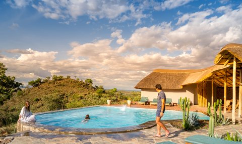 Ein Mann in kurzer Kleidung geht am Rand eines Pools, während eine Frau im Wasser entspannt und die Landschaft genießt.