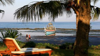 Ein ruhiger Strandblick mit einem traditionellen Boot im Wasser und einem Liegestuhl im Vordergrund, umgeben von Palmen.