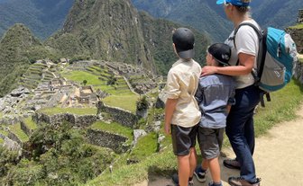 Eine Frau steht mit zwei Kindern an einem Aussichtspunkt und bewundert die Ruinen von Machu Picchu in der Ferne.
