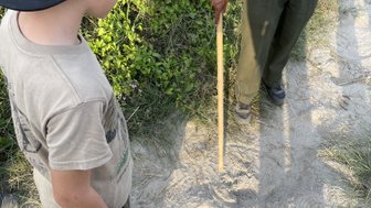 Ein Junge mit einer Kappe beobachtet aufmerksam die Fußabdrücke im Sand, während ein Erwachsener mit einem Stock daneben steht.