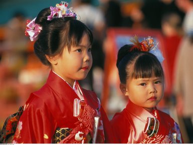 Zwei kleine Mädchen in traditioneller japanischer Kleidung, die mit Blumen im Haar an einem festlichen Ort stehen.