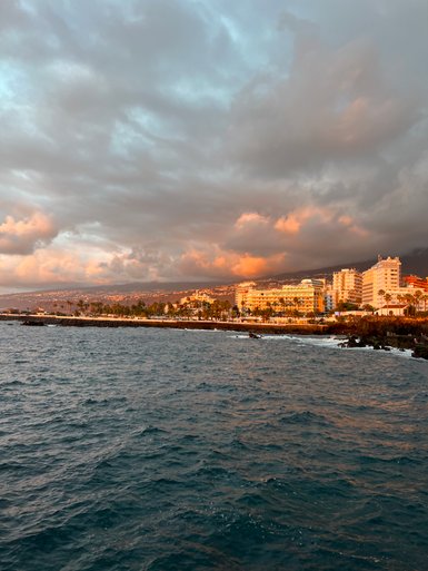 Die Küste von Puerto de la Cruz wird von sanften Wellen umspült, während die Sonne hinter den Wolken untergeht.