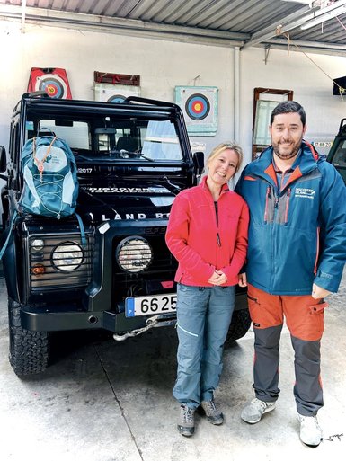 Ein Paar steht lächelnd vor einem schwarzen Geländewagen, bereit für ein Abenteuer in der Natur.