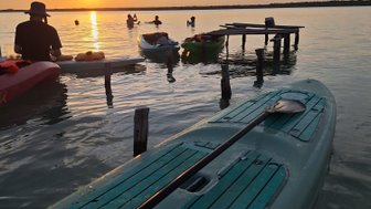 Ein ruhiger Sonnenuntergang über einem Gewässer, während Menschen in Kajaks paddeln und im Wasser schwimmen.