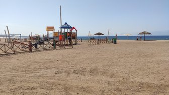 Ein bunter Spielplatz steht am Strand, umgeben von einem Zaun und mit Blick auf das ruhige Meer im Hintergrund.