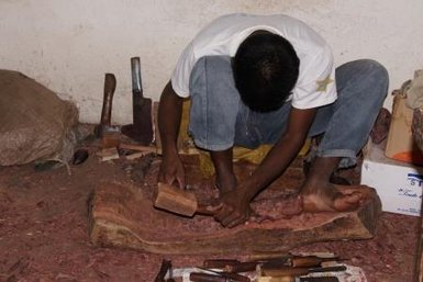 Ein Handwerker sitzt auf dem Boden und schnitzt mit einem Holzhammer an einem großen Stück Holz.