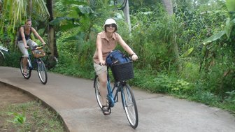 Zwei Frauen fahren mit Fahrrädern auf einem schmalen Weg, umgeben von üppigem, grünem Dschungel.