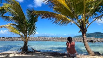 Eine Person sitzt am Strand, umgeben von Palmen, und blickt auf das ruhige Wasser und die Berge im Hintergrund.