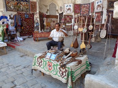 Ein Mann sitzt an einem Tisch voller traditioneller Musikinstrumente in einem lebhaften Markt mit bunten Teppichen im Hintergrund.