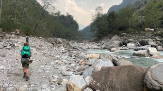 Ein Wanderer mit einem grünen Rucksack geht über einen steinigen Weg entlang eines Flusses in den Bergen Nepals.