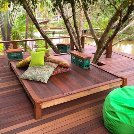 Eine einladende Holzterrasse mit bunten Kissen, umgeben von üppiger Natur und einem ruhigen Wasserlauf im Hintergrund.
