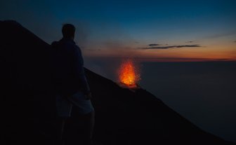 Ein Mensch steht am Rand eines Vulkans und beobachtet die beeindruckende Eruption mit glühenden Lavafontänen im Abendlicht.