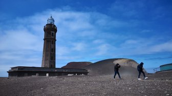 Ein alter Leuchtturm erhebt sich majestätisch über eine karge, vulkanische Landschaft unter einem klaren blauen Himmel.