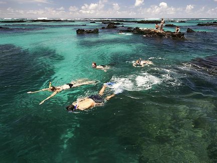 Im klaren, türkisfarbenen Wasser schwimmen mehrere Personen, während andere auf den Felsen entspannen und die Aussicht genießen.