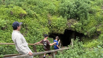 Eine Gruppe von drei Personen geht eine Holztreppe hinunter zu einem dunklen Höhleneingang, umgeben von üppigem Grün.