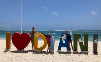 Drei Personen stehen am Strand vor bunten Buchstaben, die 'I ♥ Diani' bilden, während das Meer im Hintergrund glitzert.