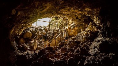 Drei Personen erkunden eine Höhle, umgeben von großen Steinen und Felsen, während Licht von einem Eingang strömt.
