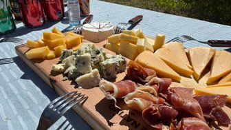 Ein rustikales Holzbrett präsentiert eine Auswahl an verschiedenen Käsesorten und Schinken, umgeben von Getränkedosen.