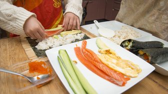 Ein Kind rollt Sushi mit frischem Lachs, Avocado und Gurke auf einem Bambusmatte, während eine andere Person im Hintergrund zuschaut.