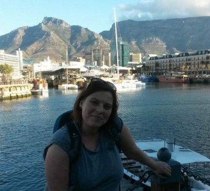 Eine Frau mit braunen Haaren lächelt am Hafen, während Boote und beeindruckende Berge im Hintergrund sichtbar sind.