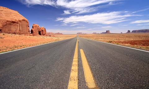 Eine endlose Straße erstreckt sich durch die weite, rote Wüstenlandschaft unter einem strahlend blauen Himmel.