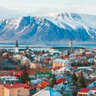 Blick auf Reykjavik mit bunten Häusern, umgeben von schneebedeckten Bergen und einem ruhigen Wasser im Hintergrund.