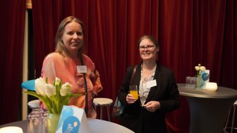 Zwei Frauen stehen an einem Tisch in einem eleganten Raum mit roten Vorhängen, lächeln und halten Getränke in der Hand.