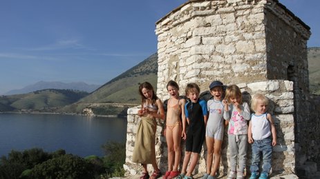 Eine Gruppe von fröhlichen Kindern steht an einem steinernen Gebäude am Wasser, umgeben von grünen Hügeln und blauen Himmel.