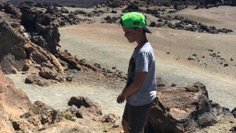 Ein Junge mit einer grünen Kappe steht auf einem felsigen Terrain, umgeben von beeindruckenden Bergen unter klarem Himmel.