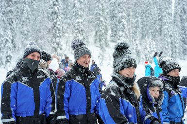 Eine Gruppe von Menschen in blauen Winterjacken lächelt im Schnee, während Schneeflocken um sie herum fallen.