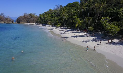 Ein sonniger Strand mit klarem Wasser, wo Menschen im Wasser schwimmen und am Ufer spazieren gehen.