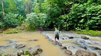 Ein Wanderer überquert einen fließenden Fluss auf einem Steinhaufen, umgeben von üppigem, grünem Dschungel.