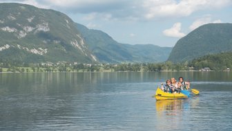 Eine Gruppe von vier Personen paddelt fröhlich in einem gelben Kajak auf dem ruhigen Bohinjsee, umgeben von grünen Bergen.