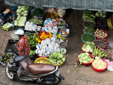 Ein belebter Marktstand in Saigon, voll mit frischem Gemüse und Obst, während eine Person in traditioneller Kleidung einkauft.