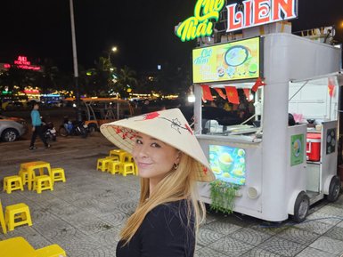 Eine lächelnde Frau trägt einen traditionellen vietnamesischen Hut und steht vor einem Straßenstand mit bunten Stühlen.