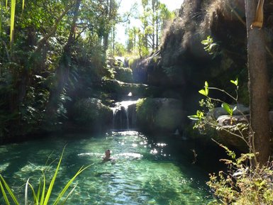 Ein klarer Naturpool umgeben von üppigem Grün und sanften Wasserfällen, in dem eine Person entspannt schwimmt.