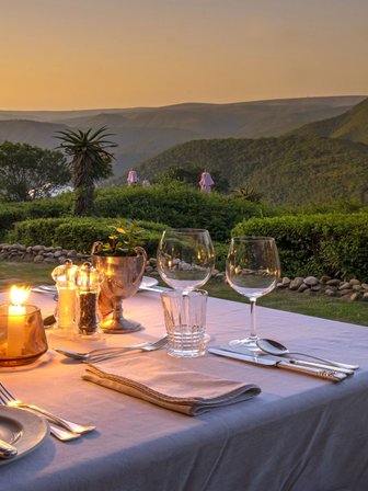 Ein romantisch gedeckter Tisch im Freien, beleuchtet von Kerzen, mit Blick auf die Berge bei Sonnenuntergang.