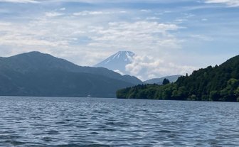 Der majestätische Mount Fuji erhebt sich über den ruhigen Wassern eines Sees, umgeben von sanften Hügeln und bewaldeten Ufern.