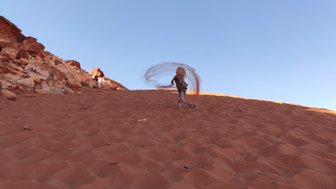 Eine Person steht auf einer sandigen Düne und wirbelt Sand in die Luft, während die Sonne am Horizont scheint.