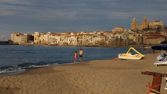 Ein Mann und ein Junge laufen am Strand entlang, während die Wellen sanft ans Ufer schlagen und die Stadt im Hintergrund sichtbar ist.