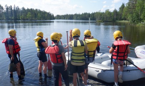 Eine Gruppe von sechs Personen in Schwimmwesten und Helmen steht am Ufer eines ruhigen Sees, bereit zum Rafting.