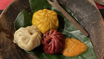 Ein rustikeller Teller mit bunten Momos, umgeben von grünen Blättern und einer kleinen Gabel aus Holz.