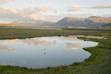 Eine ruhige Landschaft mit einem spiegelnden Gewässer, umgeben von sanften Hügeln und Bergen unter einem bewölkten Himmel.
