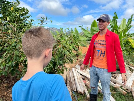 Ein Junge mit blonden Haaren steht vor einem Mann in einem roten Hemd, der in einer Kaffeeplantage spricht.