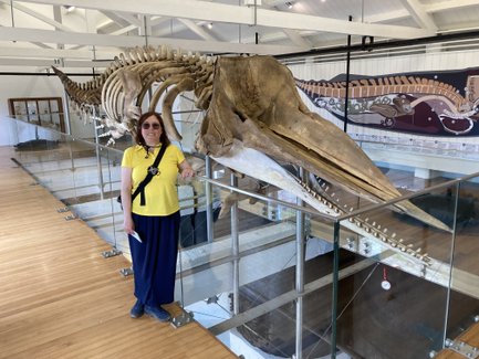 Eine Frau in einem gelben T-Shirt steht neben einem großen Walskelett in einem Museum, umgeben von Holzfußboden und Glasgeländer.