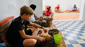 Kind hilft einheimischen Frauen beim Mahlen von Arganfrüchten zur Ölgewinnung in Essaouira – Marokko Familienreise