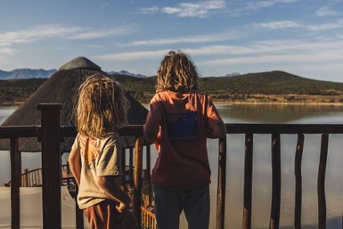 Zwei Kinder mit lockigem Haar stehen an einem Geländer und blicken auf einen ruhigen See und die umliegenden Berge.