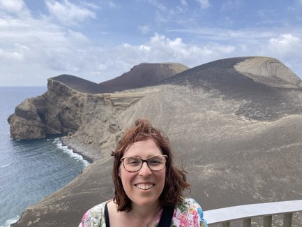 Eine Frau mit lockigem, braunem Haar lächelt vor einer beeindruckenden vulkanischen Landschaft am Meer.