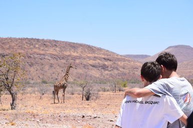 Zwei Kinder beobachten gespannt eine Giraffe - Namibia mit Jugendlichen