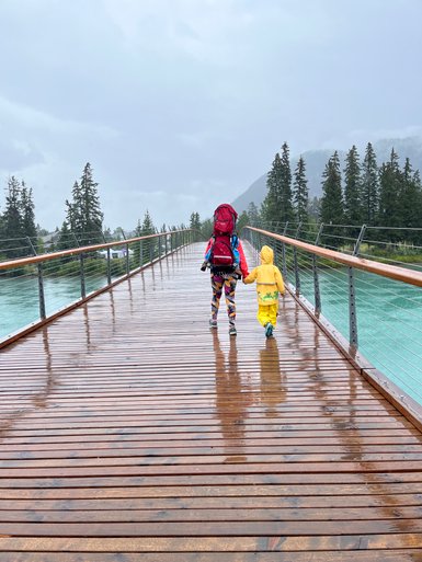 Eine Person mit einem großen Rucksack und ein Kind in gelber Regenkleidung gehen Hand in Hand über eine Holzbrücke.